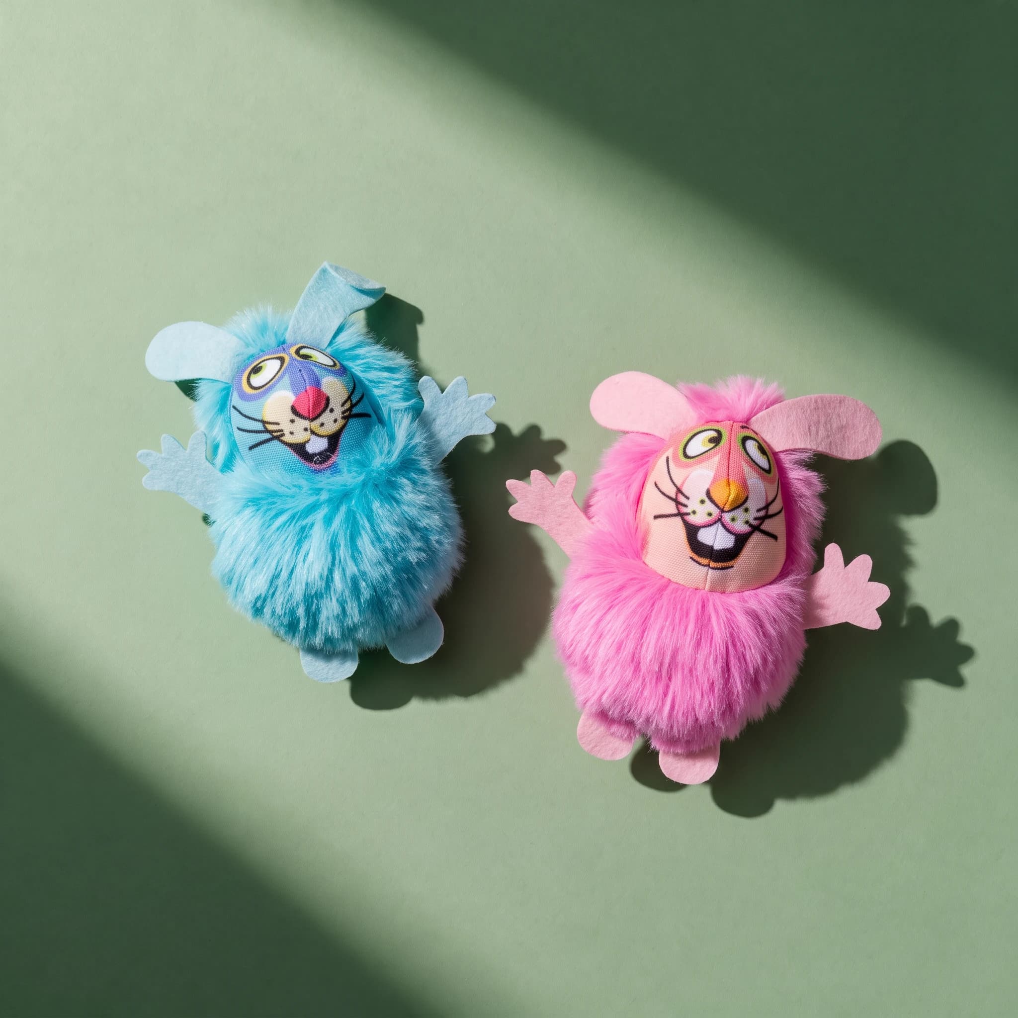 Two Fat Cat Fluff Bunnies toys shown together from above, one in blue fur and one in pink fur, each with felt ears, felt paws, and a printed cartoon bunny face, against a sage green background.