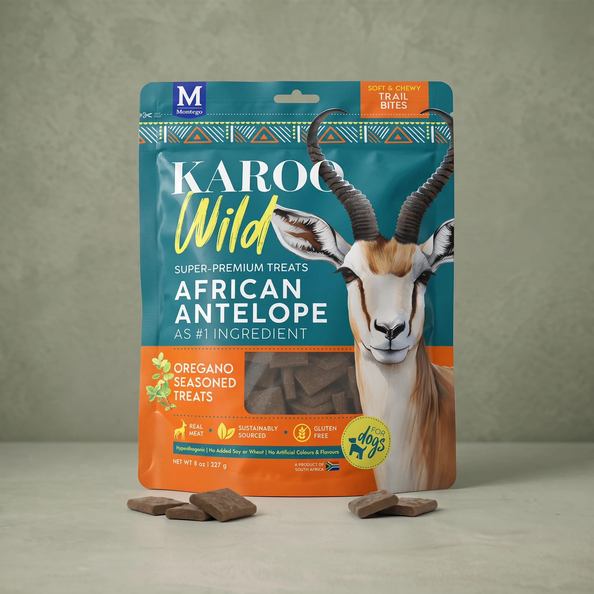 The same teal and orange Karoo Wild Oregano Trail Bites pouch standing upright against a plain sage green background, with small square dark brown treats scattered at its base.