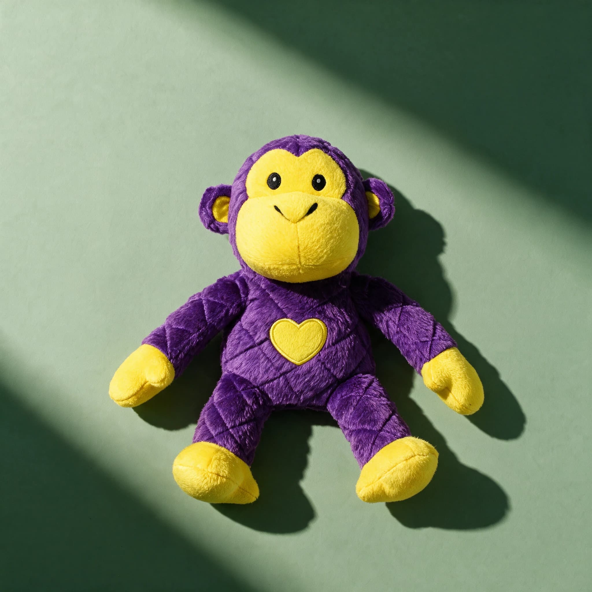 Close-up shot of the Mighty Toys Safari Monkey plush dog toy alone, lying flat with its purple quilted diamond-stitched body and limbs, yellow face, ears, hands and feet, black button eyes, a stitched smile, and a yellow embroidered heart patch on its chest, against a sage green background with a strong diagonal shadow.