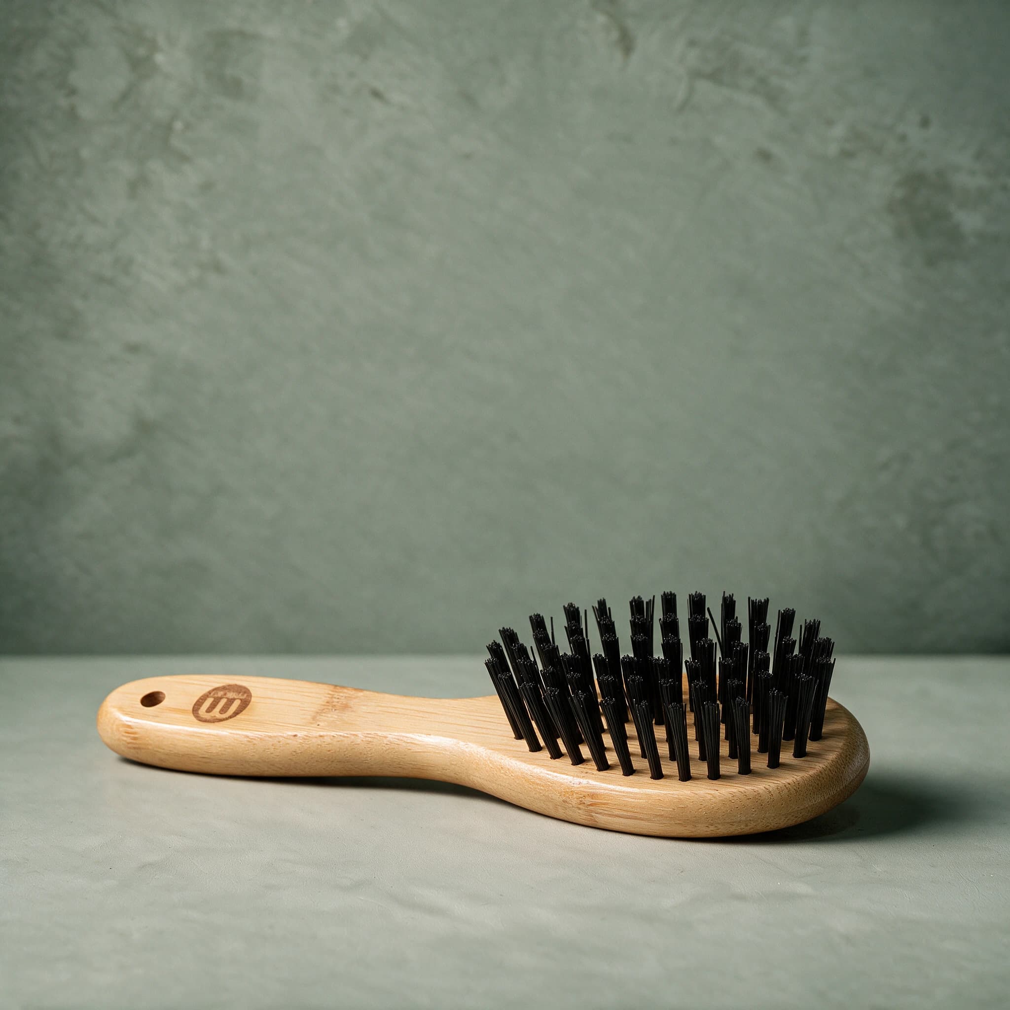 Mikki Bamboo Bristle Brush with an oval bamboo head set with nylon bristles, shown on a sage green background.