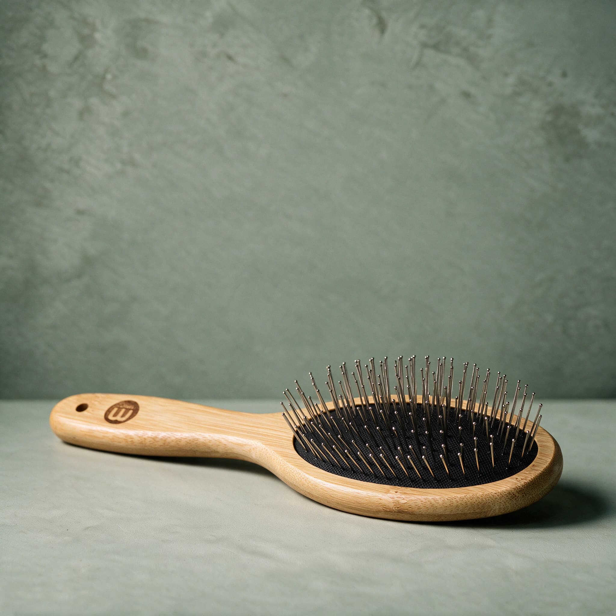 Mikki Bamboo Pin Brush with an oval bamboo head set with rounded stainless steel pins, shown on a sage green background.