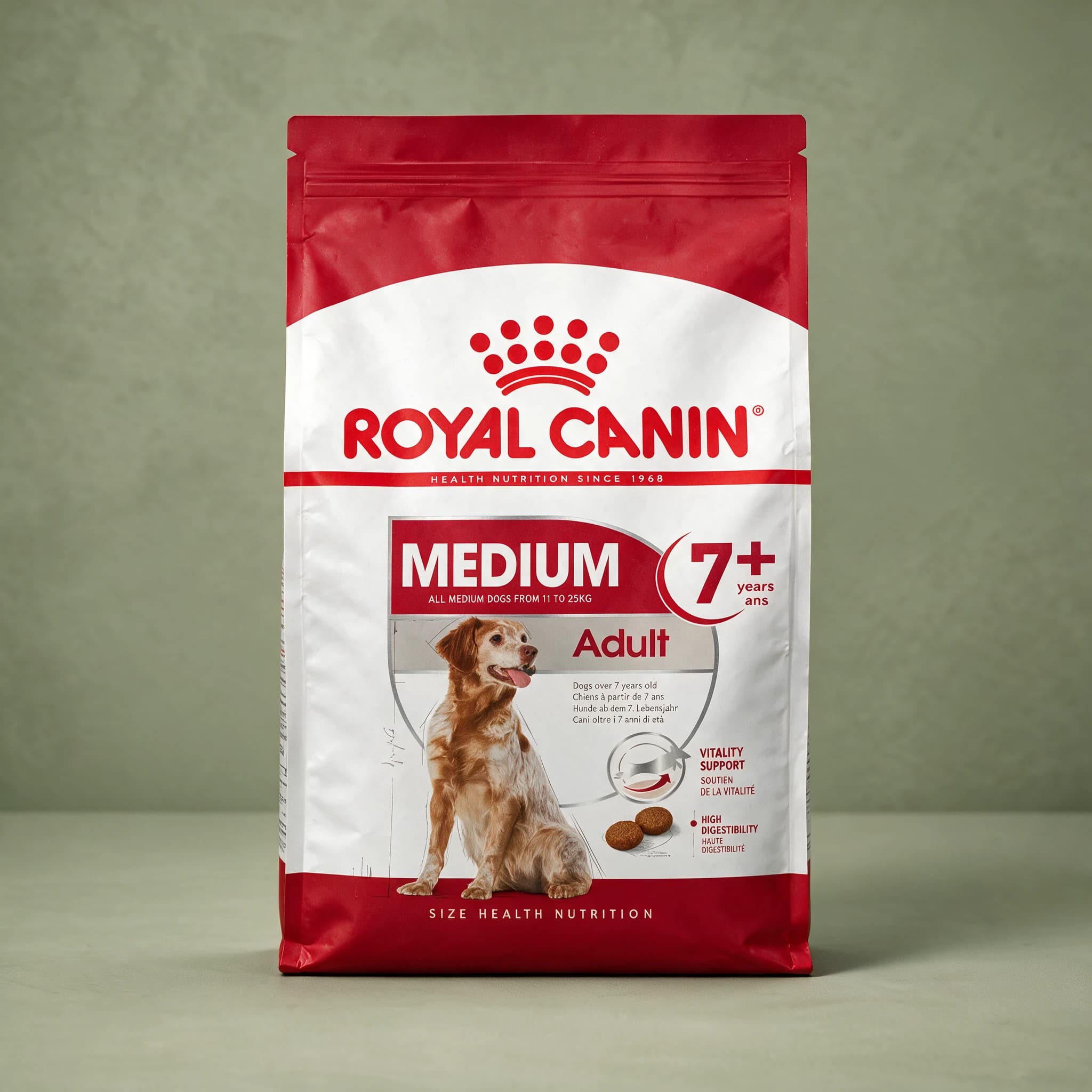 A grey and white Royal Canin Medium Adult 7+ dry dog food bag with a mature medium-breed dog illustrated on the front, photographed against a plain sage green studio background.