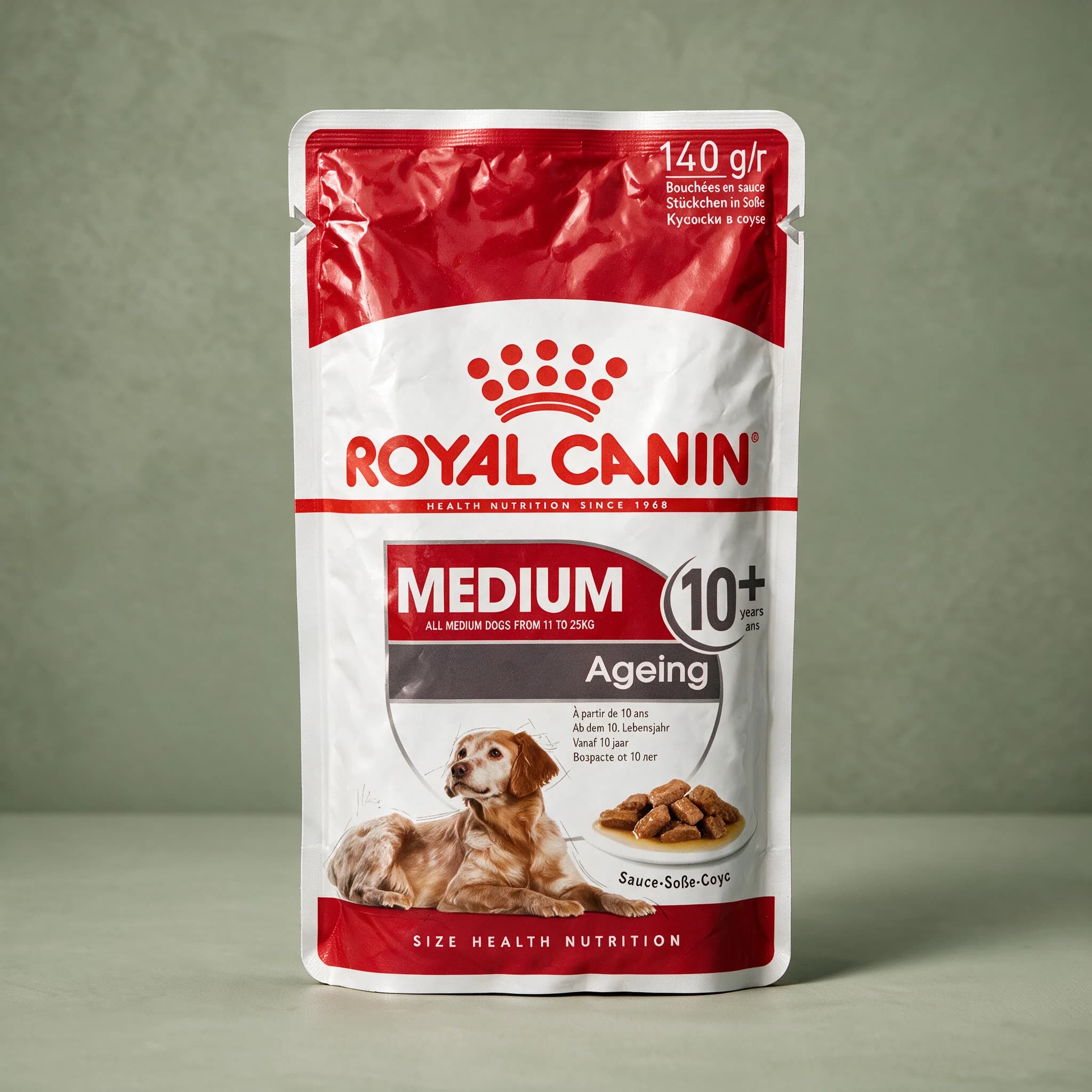 A Royal Canin Medium Ageing 10+ wet dog food pouch with the red crown logo and a senior dog illustration, standing upright against a plain sage green studio background.