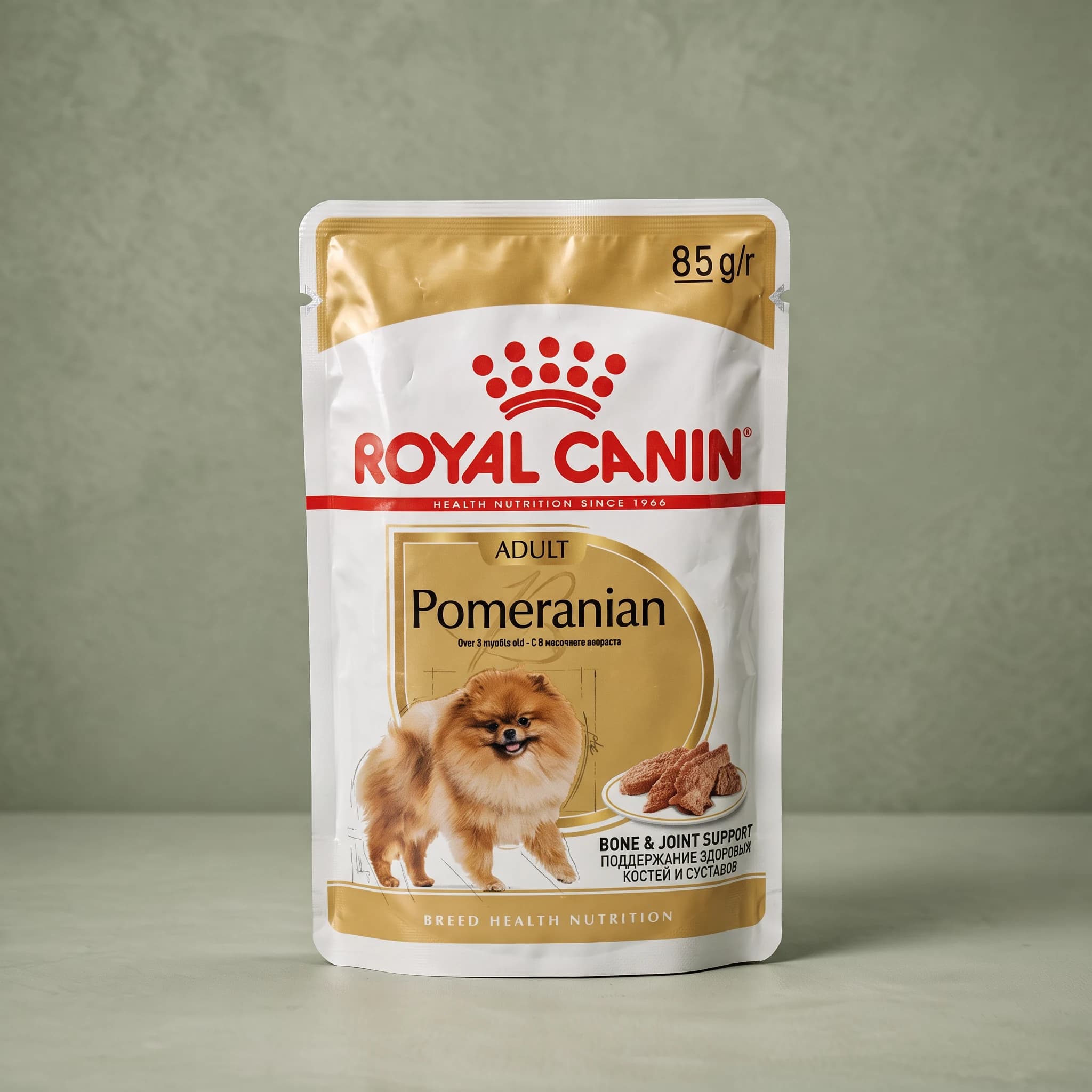 A gold and white Royal Canin "Pomeranian Adult" wet dog food pouch with the red crown logo, an illustration of a fluffy cream Pomeranian, a "Bone & Joint Support" callout, and an "85g/r" sliced-loaf label, standing upright against a plain sage green studio background.