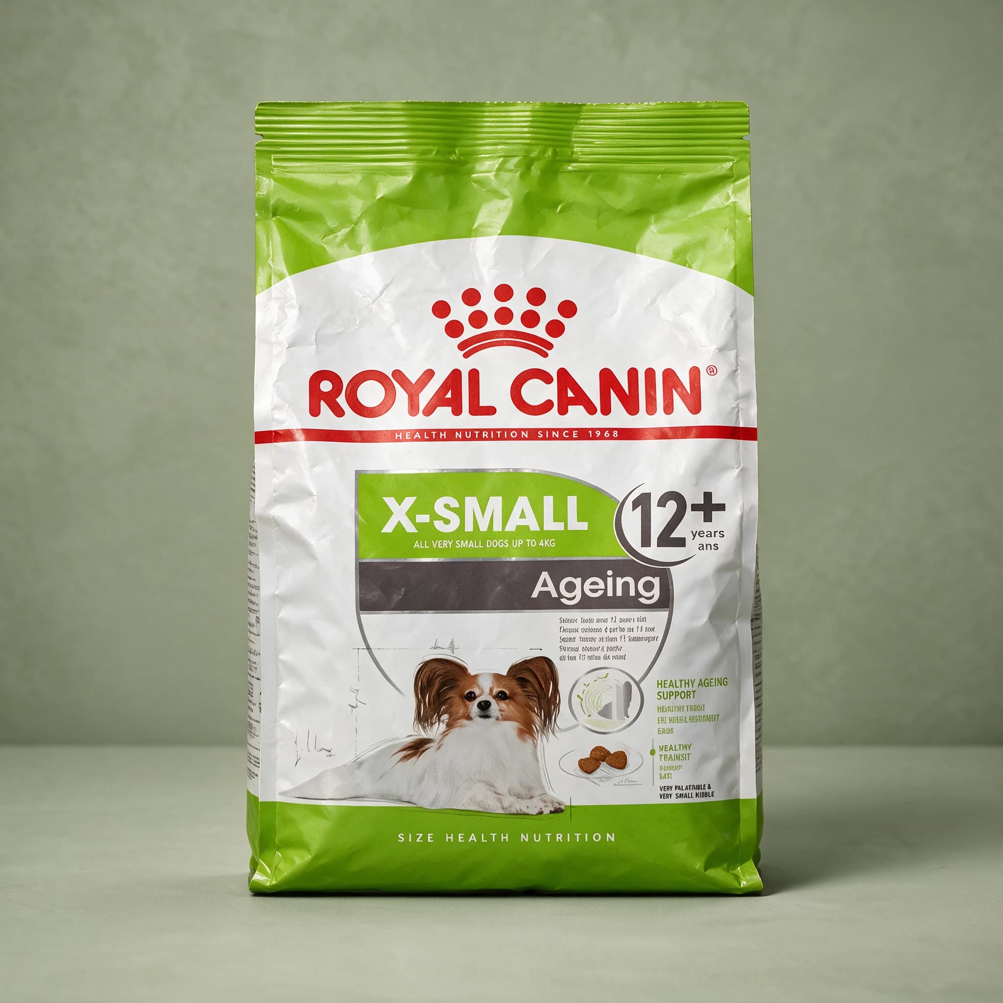 A grey and white Royal Canin bag labeled "Ageing X-Small" for senior toy-breed dogs, standing upright against a sage green studio background with the red crown logo prominent.