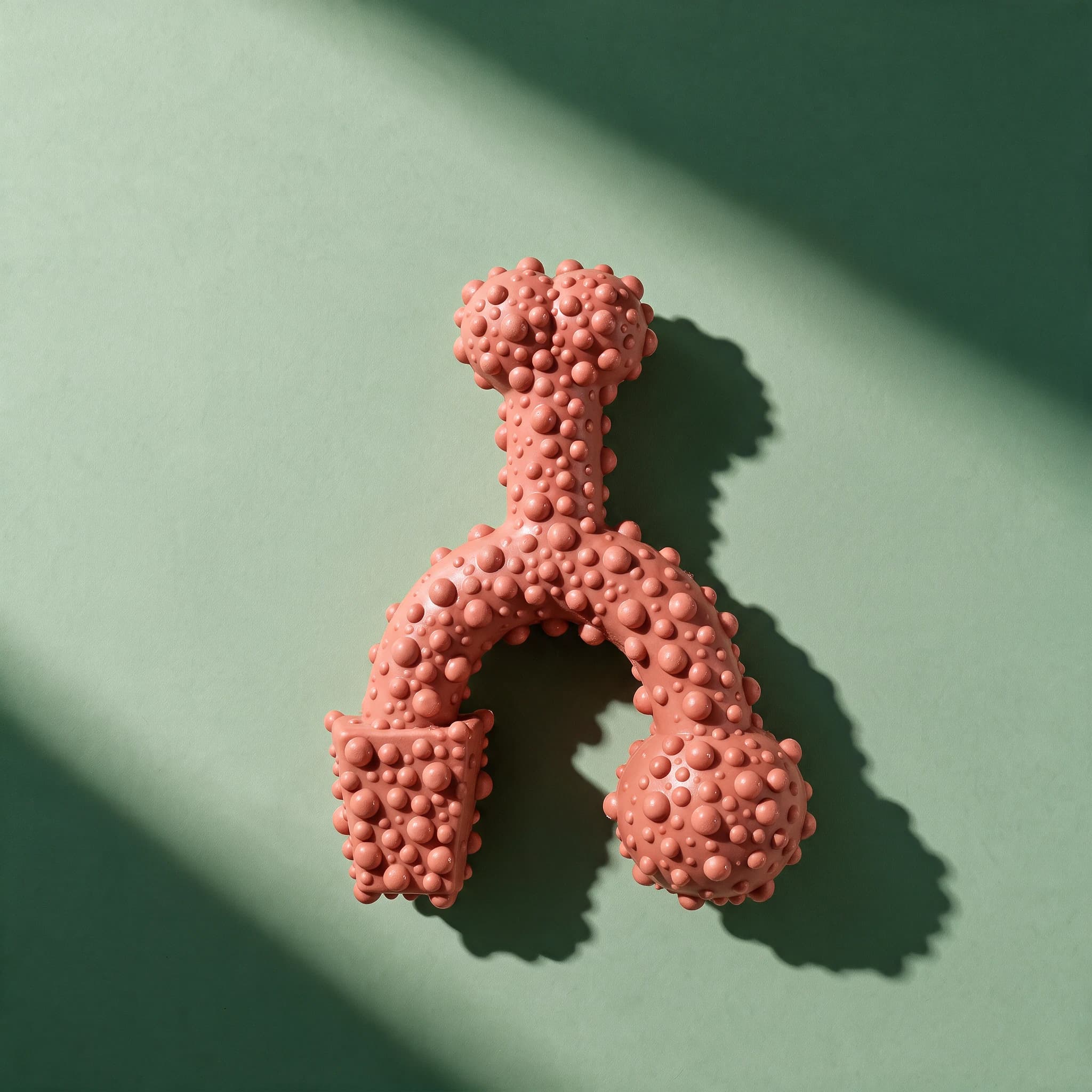 A close-up of the pink Tasty Bone Boerewors chew toy, highlighting its forked, wishbone-like shape and dense nubby texture, against a green background.