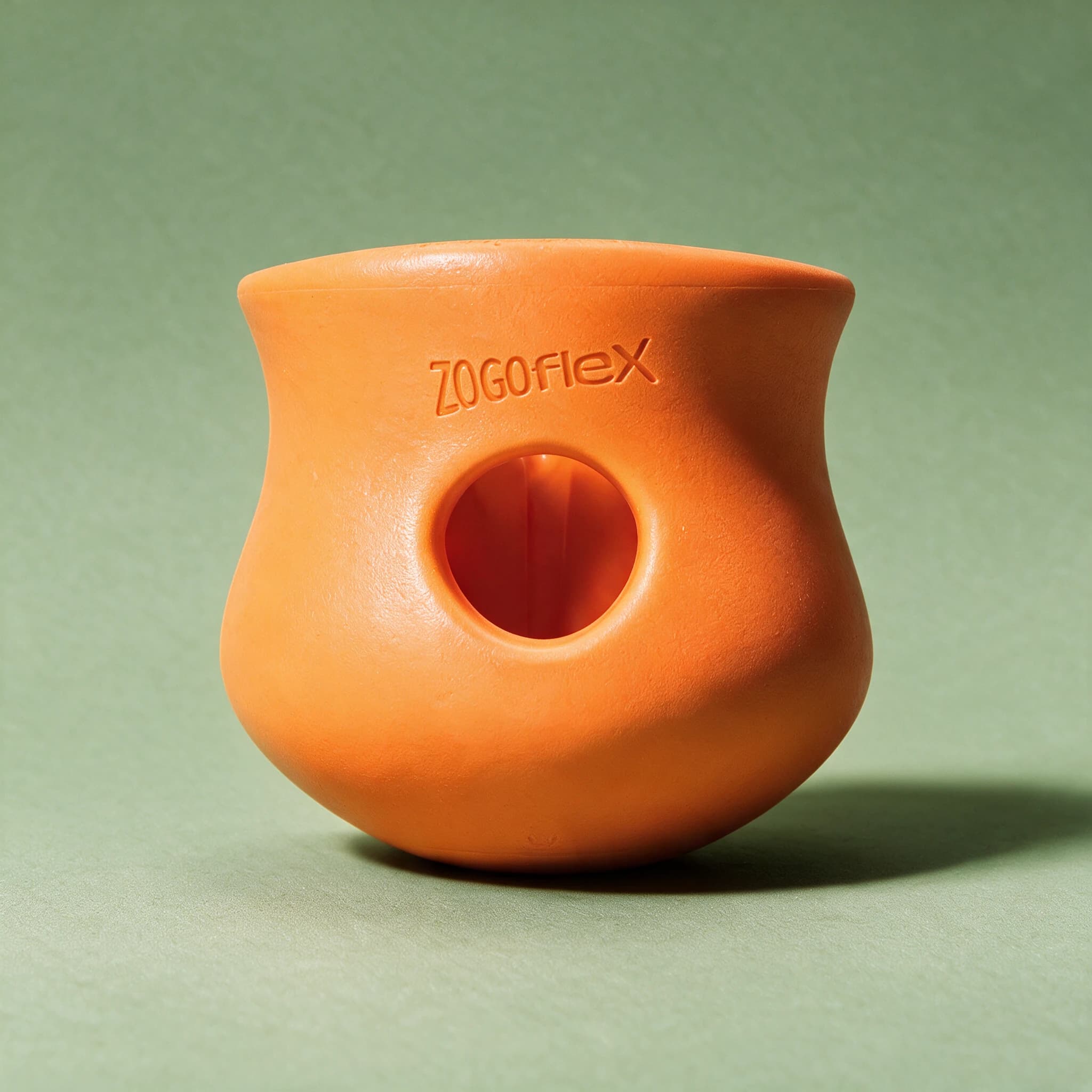 Close-up studio shot of the orange West Paw Zogoflex Toppl treat toy, showing its wide-mouthed, curved cup shape with a hollow centre hole and embossed "Zogoflex" logo, against a sage green background.