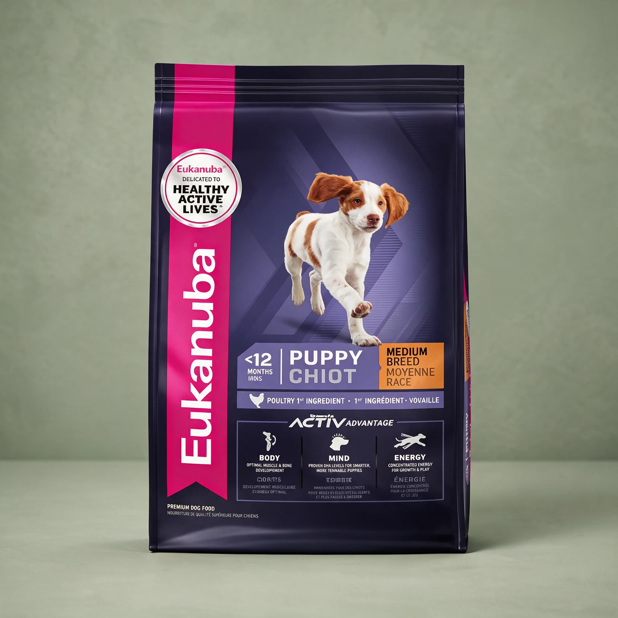 The purple and navy Eukanuba Active Advantage Medium Breed Puppy dry dog food bag with a pink ribbon and "Healthy Active Lives" seal, featuring a running brown-and-white Brittany-type puppy and a "Chicken 1st Ingredient" banner, photographed against a sage green studio background.